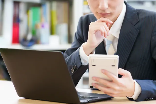 スーツ姿の男性が電卓を持ちながらノートパソコンの前で考え込んでいる様子